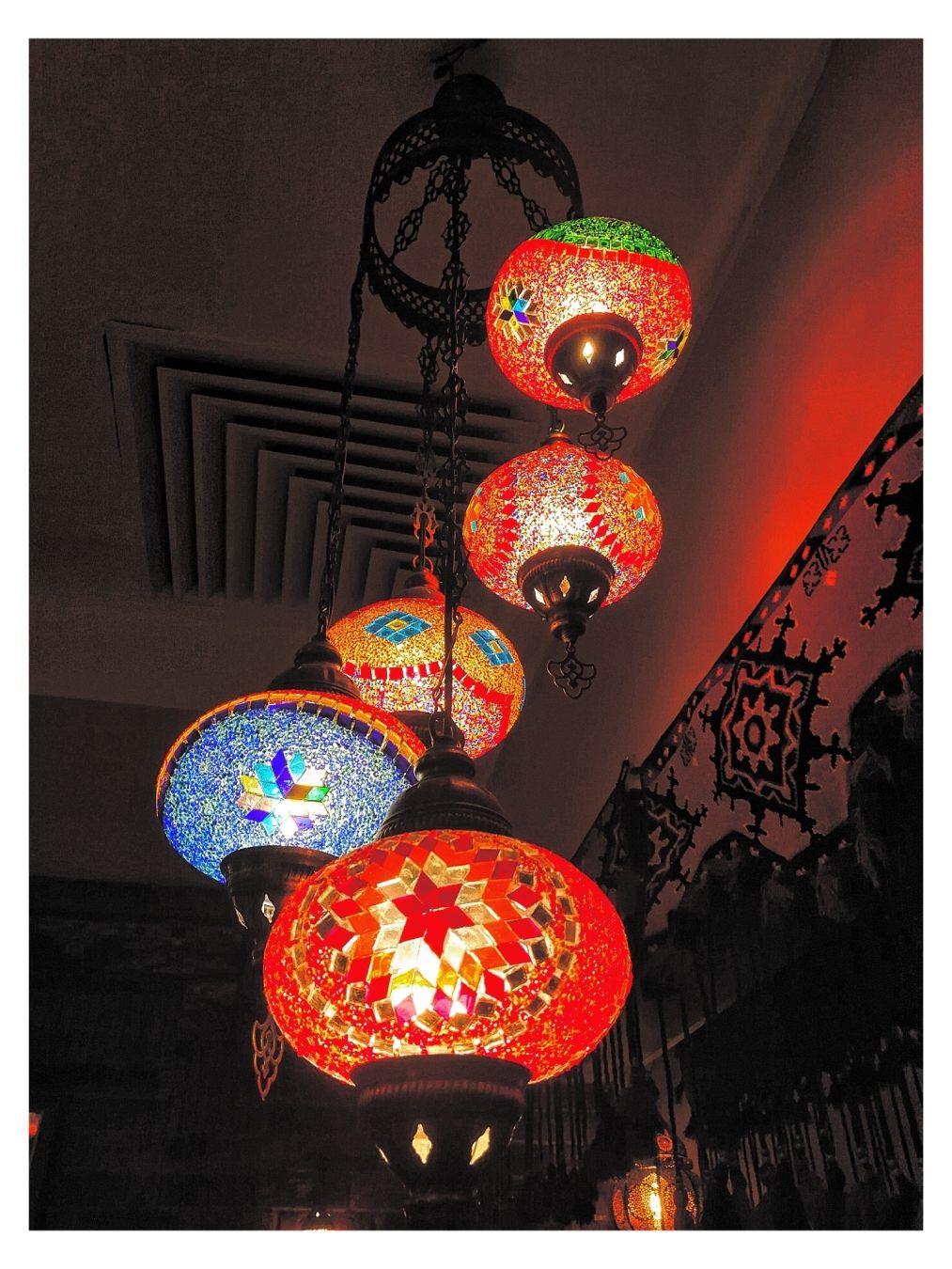 Uno scatto di un lampadario multilivello con diverse lampade rotonde a mosaico. Le lampade sono di vari colori, tra cui rosso, arancione, verde e blu, con una luce calda e luminosa che emana da ognuno. Il lampadario è sospeso dal soffitto e una parte di una parete modellata è visibile sullo sfondo.