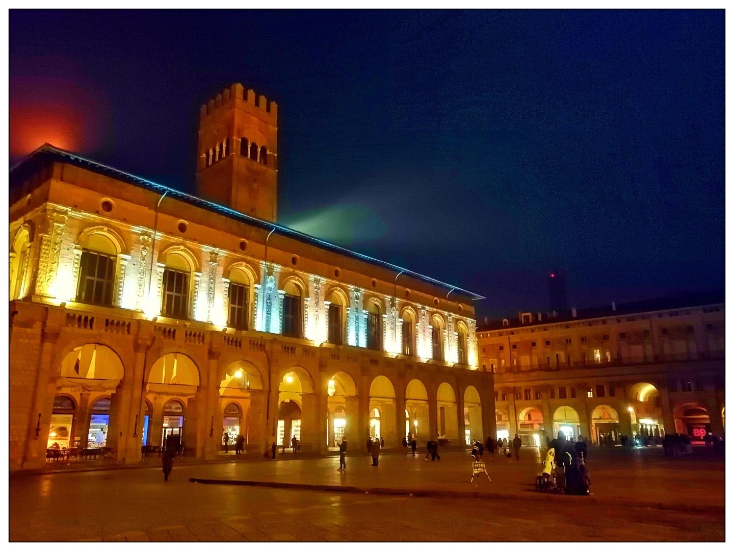 Una visione notturna di un grande palazzo illuminato (palce) con sentieri ad arco al piano terra. Una torre alta e quadrata sorge dal tetto del palazzo. In una grande piazza aperta, è visibile un secondo edificio più breve con un'architettura simile. Diverse persone camminano in piazza. Il cielo è buio.