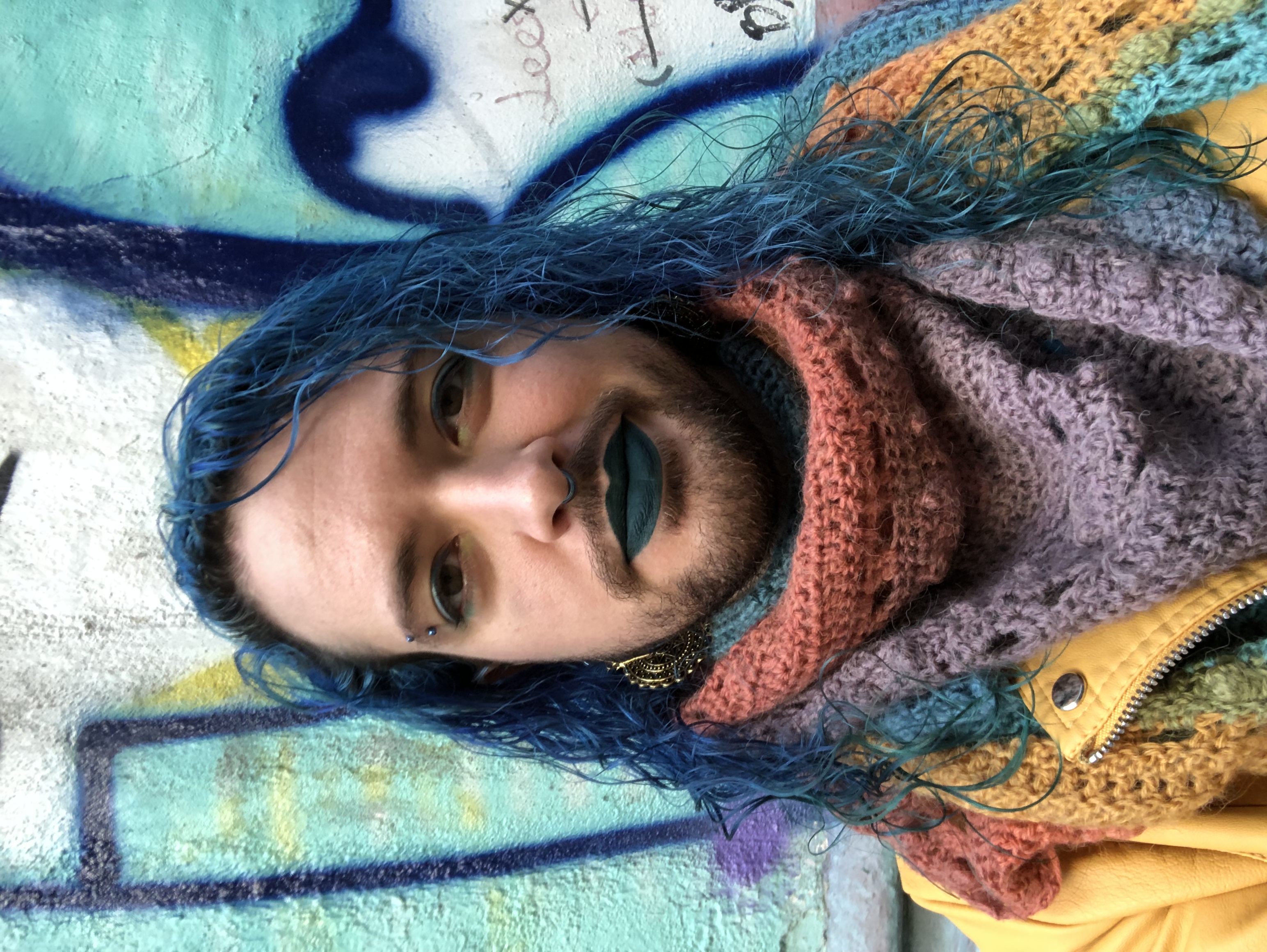 Ártemis, a white queer with curly blue hair and a beard. They’re wearing eyeshadow and lipstick in various shades of blue and turquoise, a bright yellow pleather jacket, and a crocheted rainbow scarf.