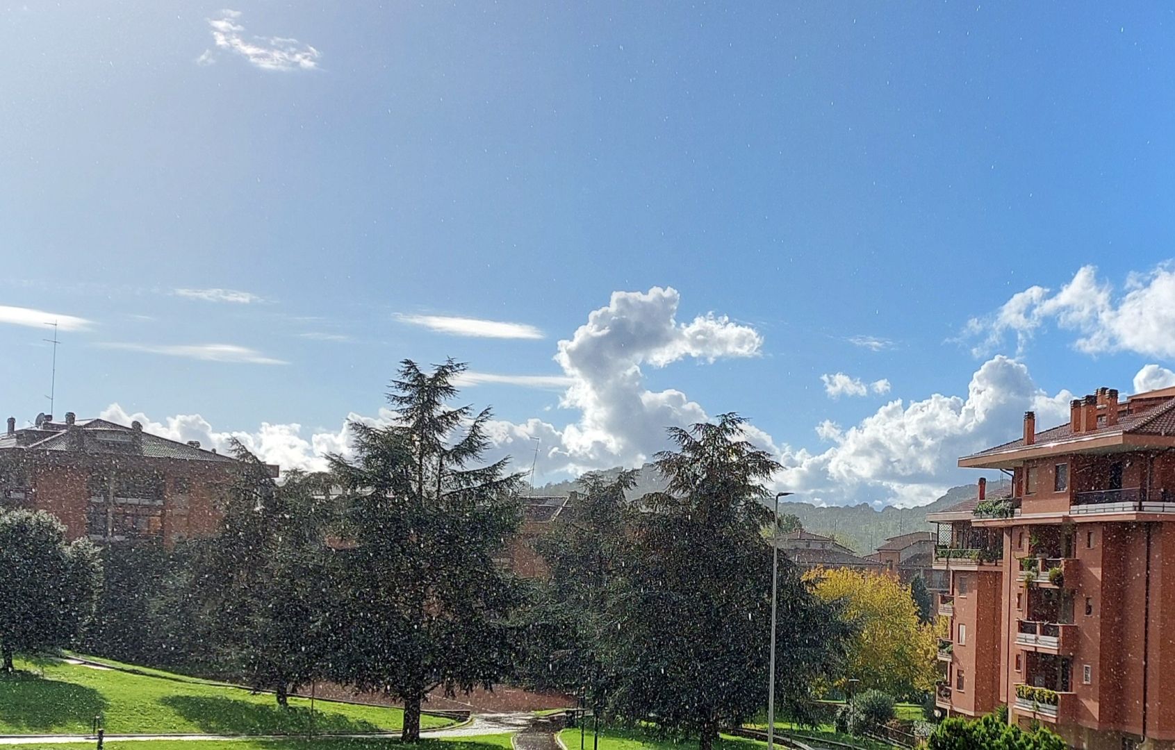 Parsaggio urbano, vista su un parco, a destra i palazzi, in alto un cielo quasi sgombro di nubi, il sole illumina tutto mentre piove.
Cityscape, view of a park, buildings on the right, a sky almost free of clouds above, the sun illuminates everything while it rains.