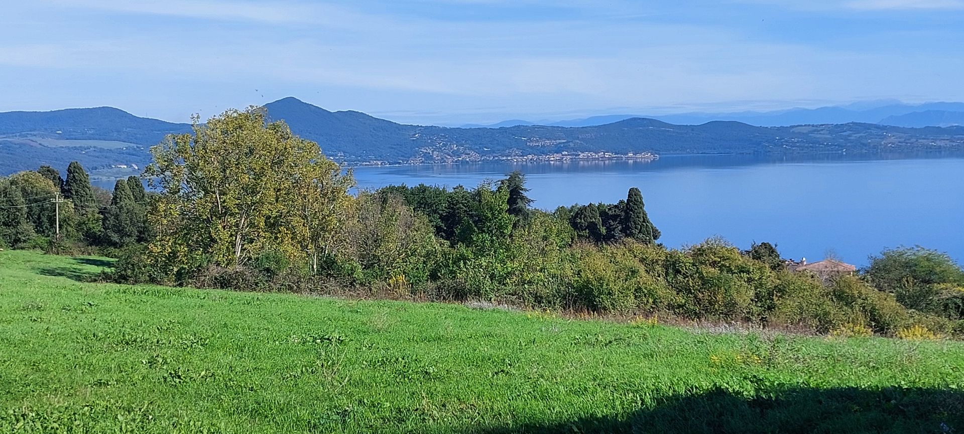 Parziale vista del lago, in primo piano un prato verde, poi il lago e le colline sotto un cielo limpido.
Partial view of the lake, in the foreground a green meadow, then the lake and the hills under a clear sky.