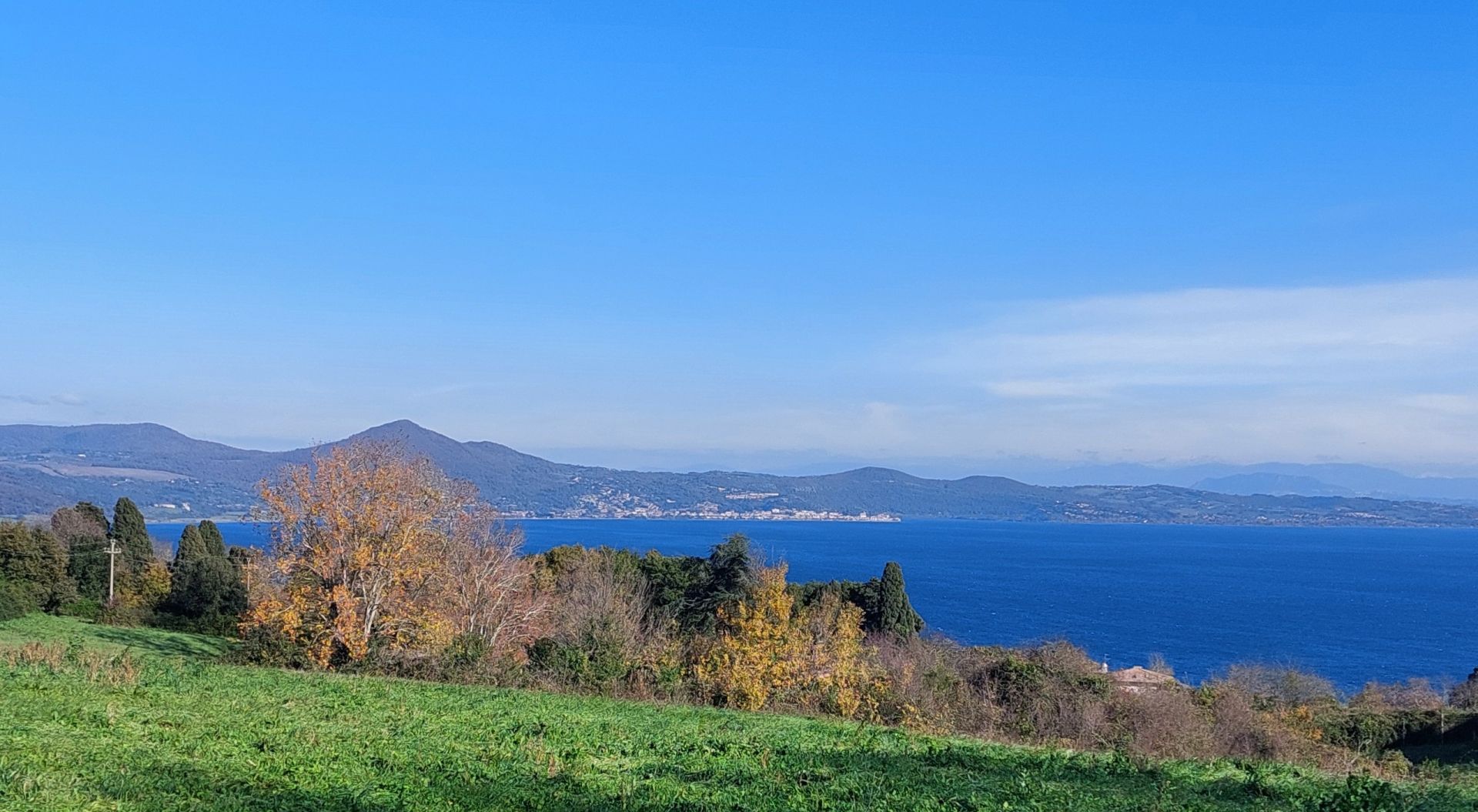 Solita immagine del lago in una assolata giornata di autunno inoltrato. In alto il cielo sereno, al centro una parte del lago, in basso alcuni alberi e un prato verde. 
Usual image of the lake on a sunny day in late autumn. Above the clear sky, in the center a part of the lake, below some trees and a green meadow.