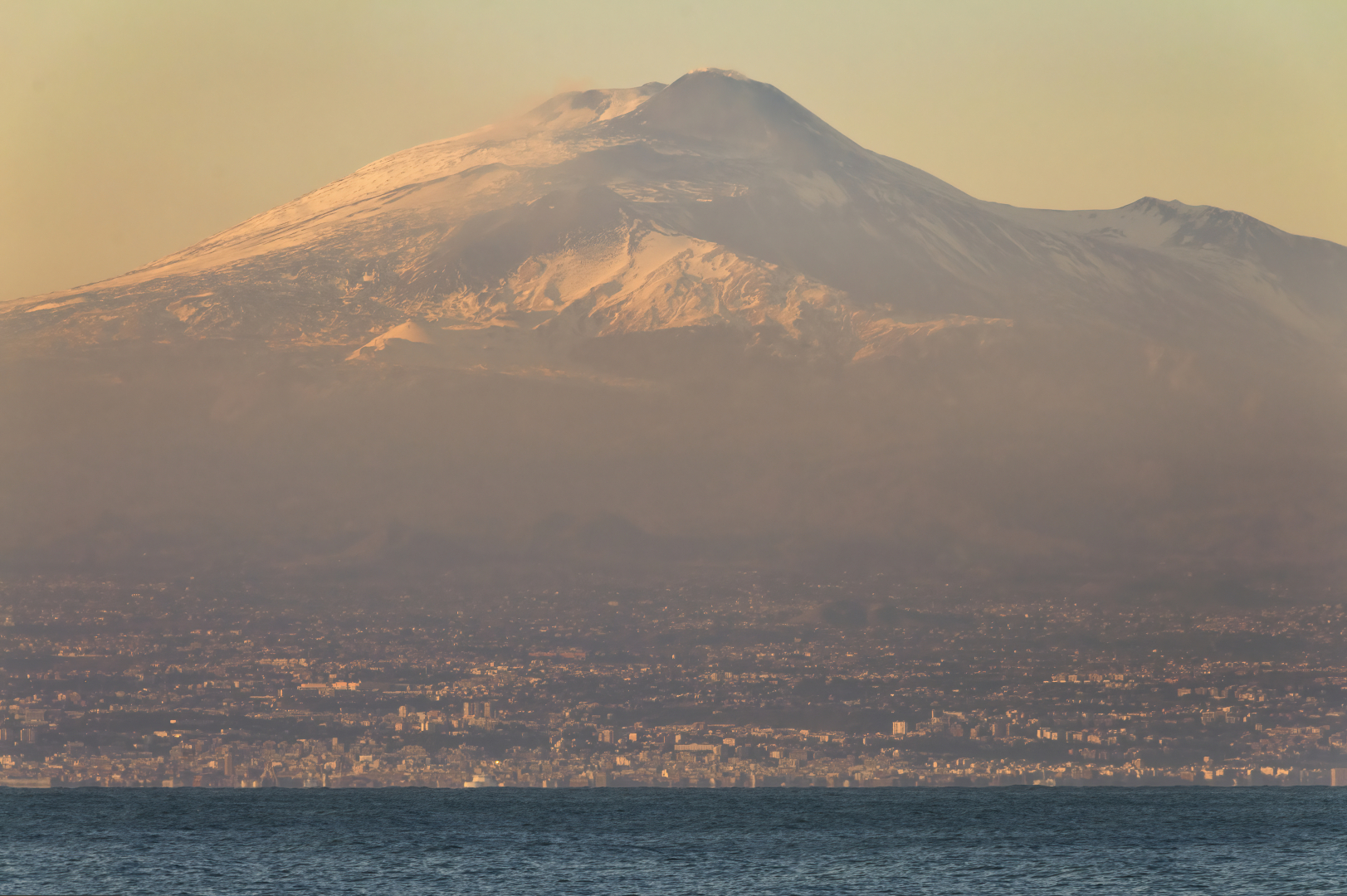 La città di Catania fotografata da lontano, al tramonto, con l'Etna che incombe su di lei.