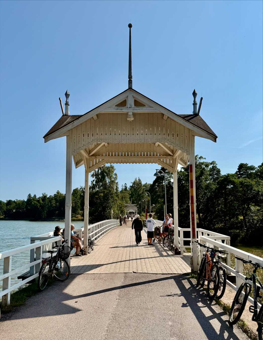 The historic white wooden bridge leading to Seurasaari Island in Helsinki, Finland, stretches across calm Baltic waters beneath a clear blue summer sky. Visitors walk and cycle across the bridge while bicycles rest along the railings, creating a welcoming scene that marks the entrance to the Seurasaari Open-Air Museum and its tranquil island setting.