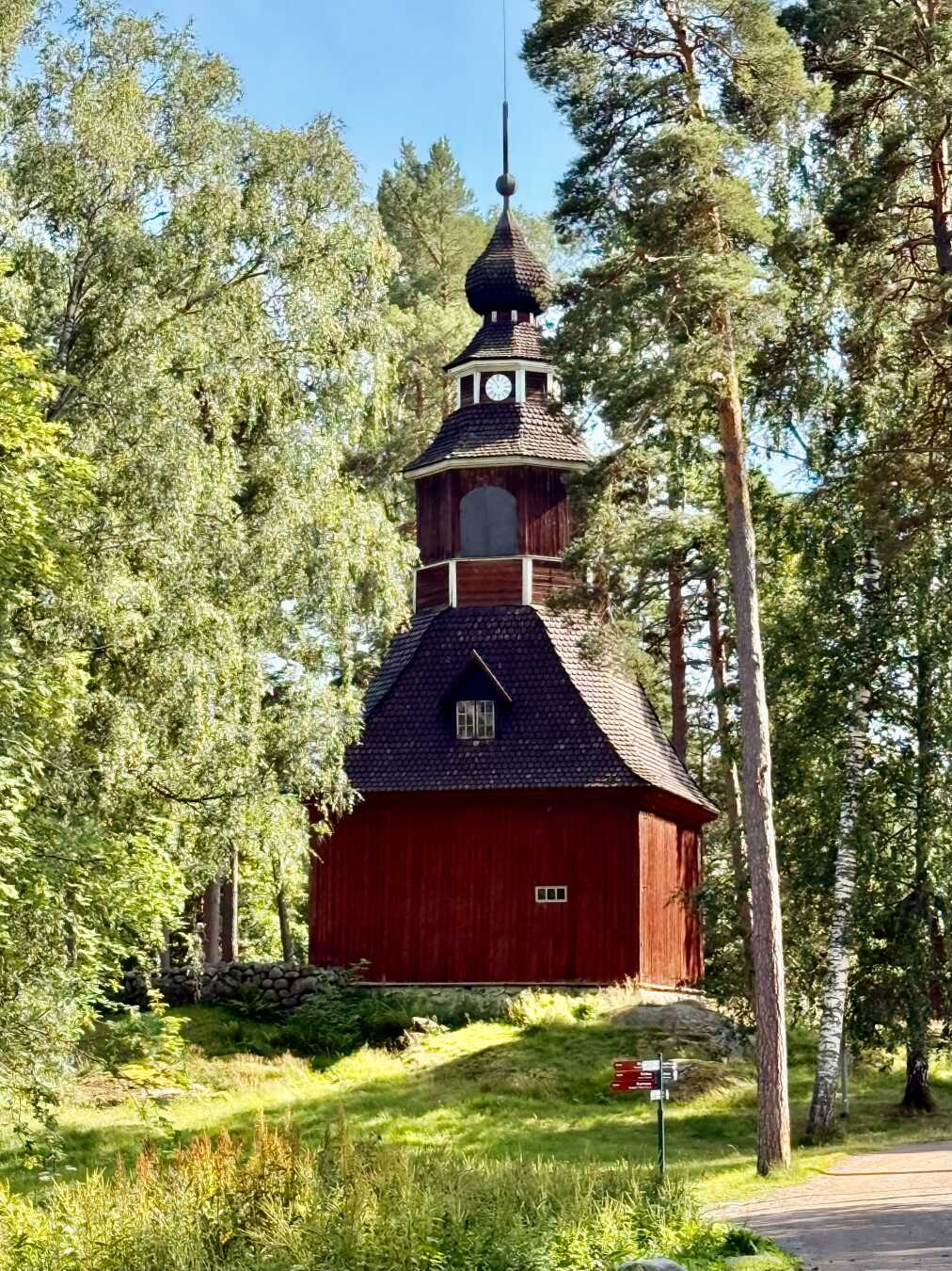 Traditional red wooden church nestled among tall pine and birch trees on Seurasaari Island in Helsinki, Finland. The historic bell tower features a dark shingled roof, a small clock, and an onion-shaped spire rising above the forest canopy. Warm afternoon sunlight filters through the trees, illuminating the peaceful woodland setting and highlighting the church’s distinctive Nordic architecture.