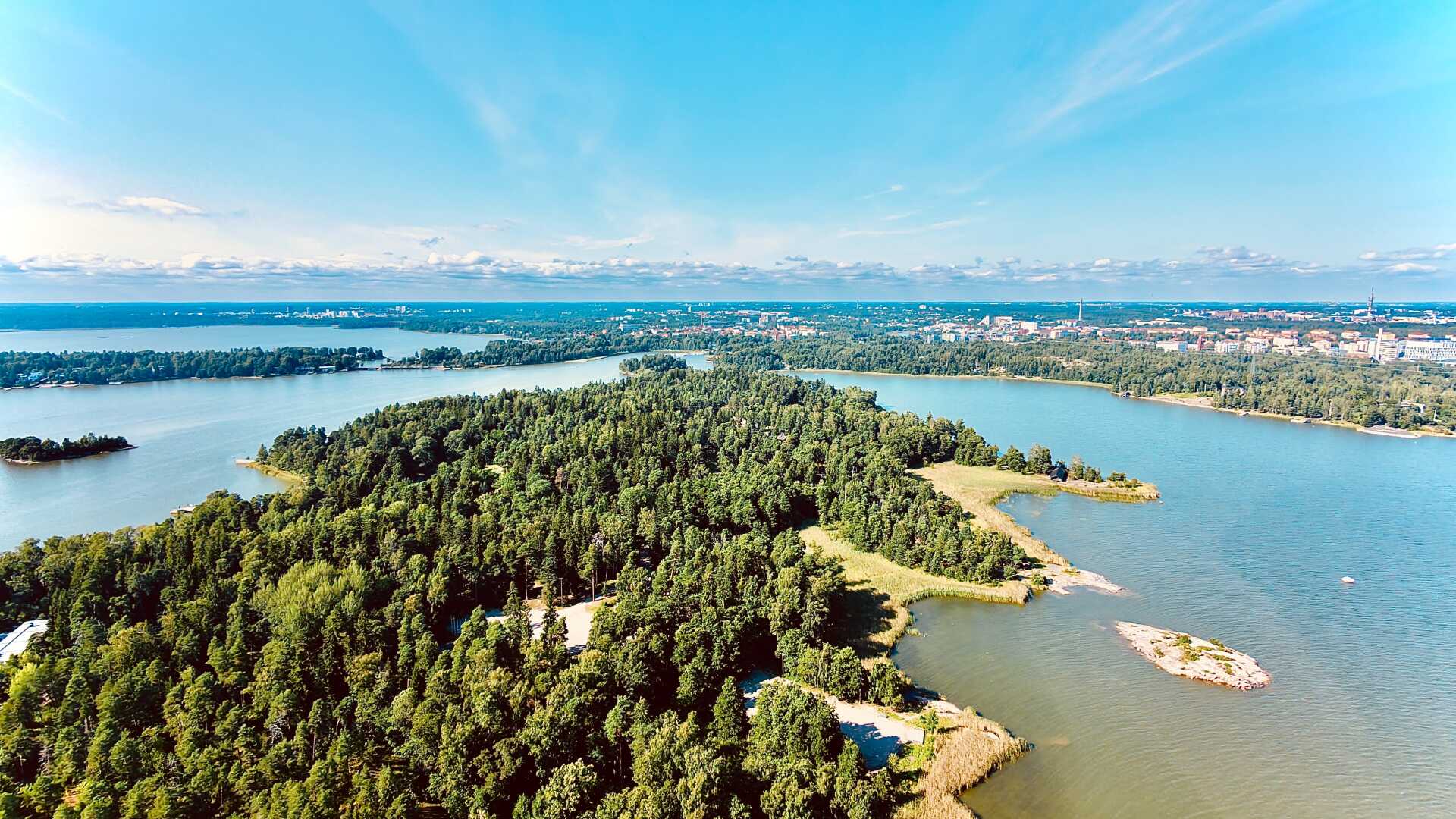 Aerial view of Seurasaari, a forested island in Helsinki, Finland, surrounded by the calm waters of the Baltic Sea. Rocky shorelines and small coves frame the lush green landscape, while the Helsinki skyline stretches across the horizon under a clear blue sky with scattered clouds. The wide-angle perspective highlights the contrast between the island’s natural scenery and the nearby urban coastline.
