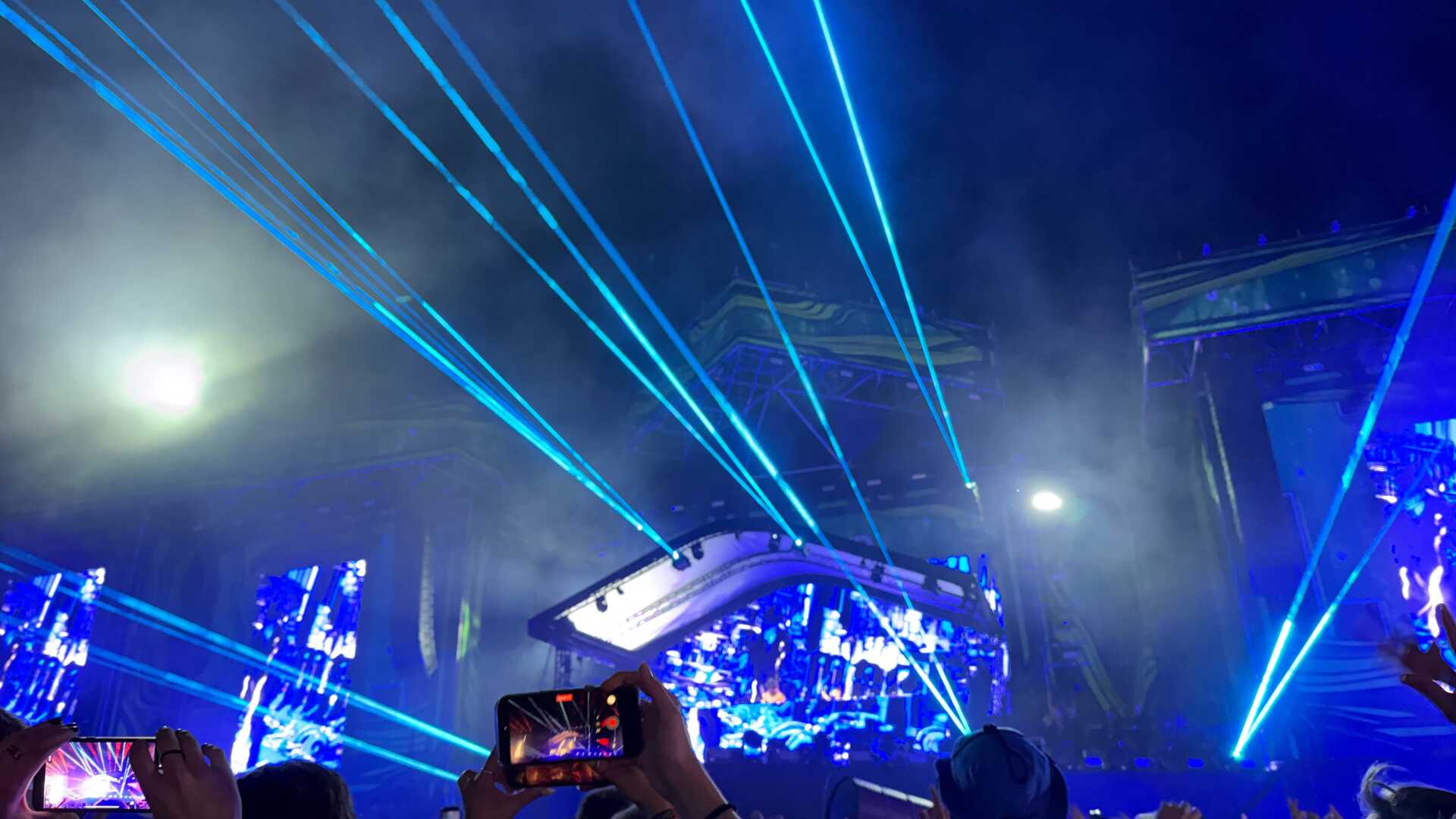 Audience members watch a DJ perform at Weekend Festival in Helsinki, Finland, as vivid blue laser beams sweep across the night sky above the main stage. Spectators hold up their phones to capture the immersive electronic music show.