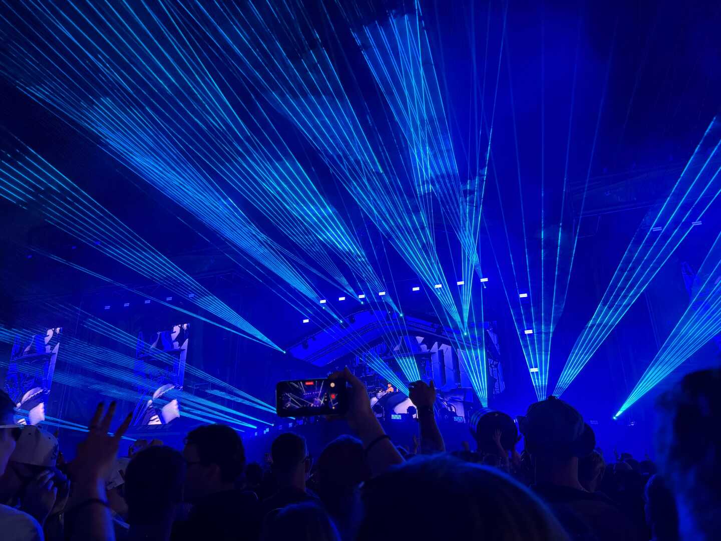 Audience members watch a DJ perform at Weekend Festival in Helsinki, Finland, as vivid blue laser beams sweep across the night sky above the main stage. Spectators hold up their phones to capture the immersive electronic music show.