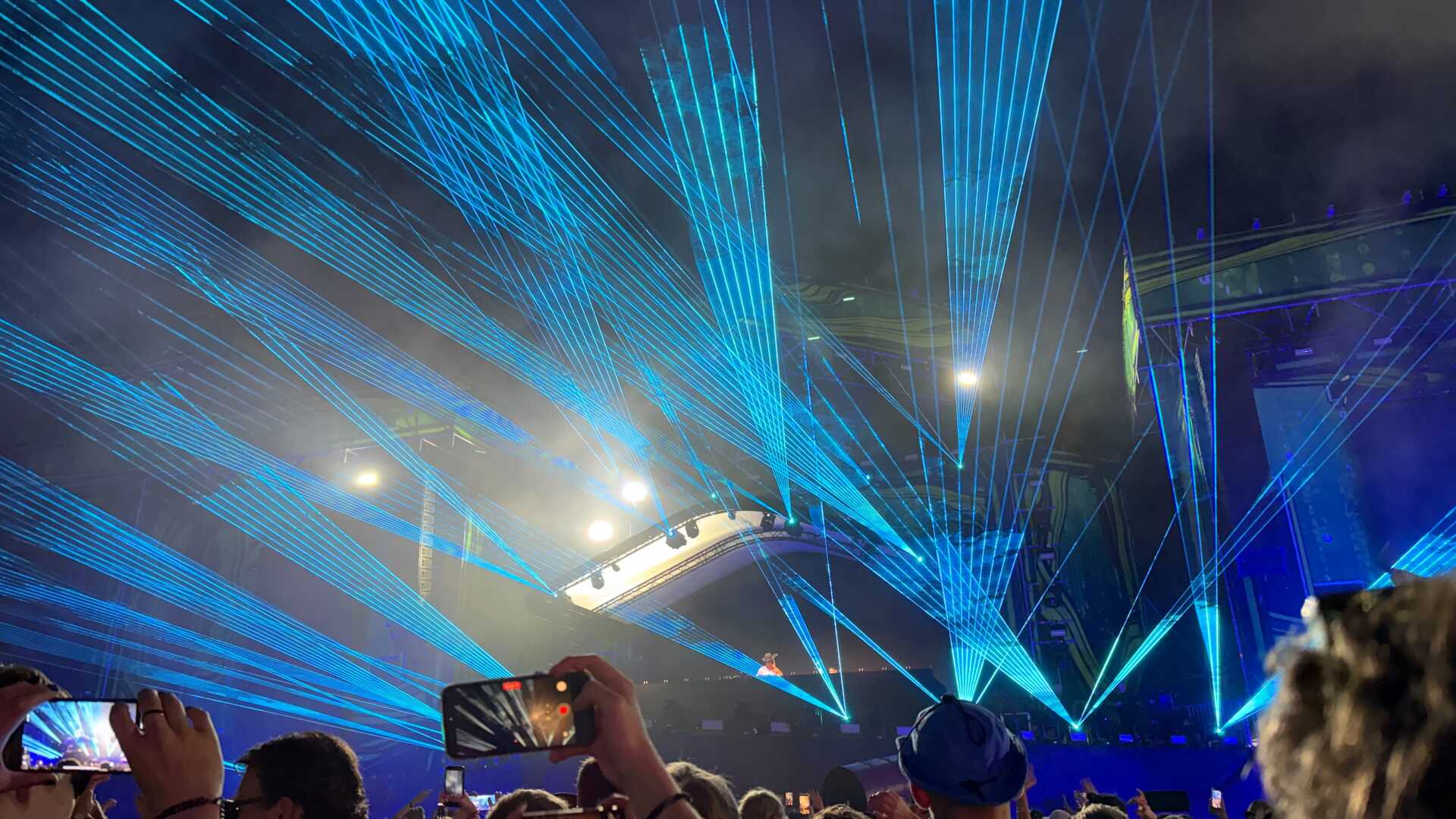 Audience members watch a DJ perform at Weekend Festival in Helsinki, Finland, as vivid blue laser beams sweep across the night sky above the main stage. Spectators hold up their phones to capture the immersive electronic music show.