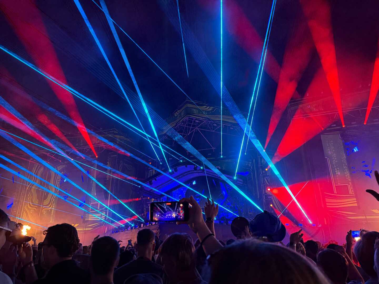 Audience members watch a DJ perform at Weekend Festival in Helsinki, Finland, as vivid blue laser beams sweep across the night sky above the main stage. Spectators hold up their phones to capture the immersive electronic music show.