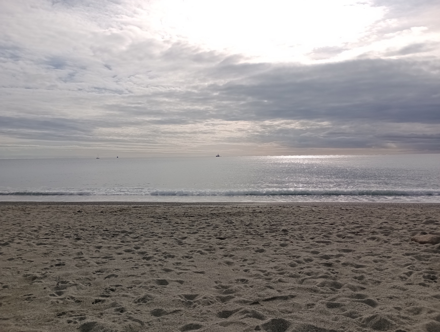 Nella foto al centro si può vedere il mare calmo del mattino con il riflesso del sole. Nella fascia inferiore la spiaggia e in quella superiore il cielo nuvoloso con alcuni raggi di sole.