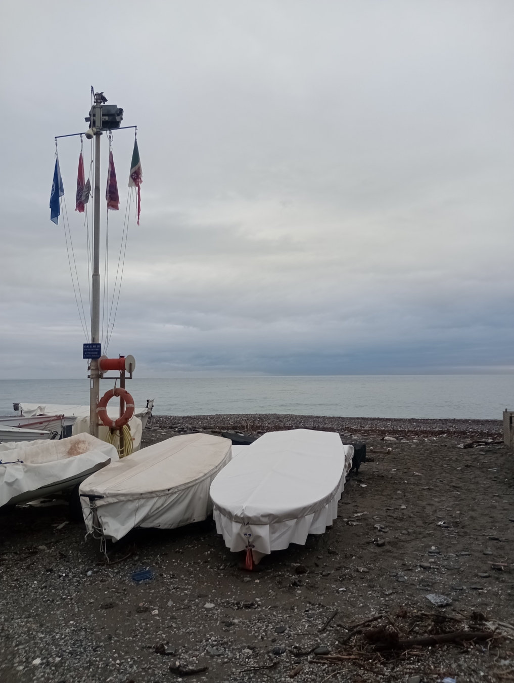 Nell'immagine si vedono due barche di pescatori (gozzi) in spiaggia. Le barche sono tirate a riva e coperte. C'è un palo con in cima un riflettore e su questo palo prendono 4 bandiere. 
In lontananza, nella fascia centrale, c'è il mare e sopra il cielo nuvoloso.