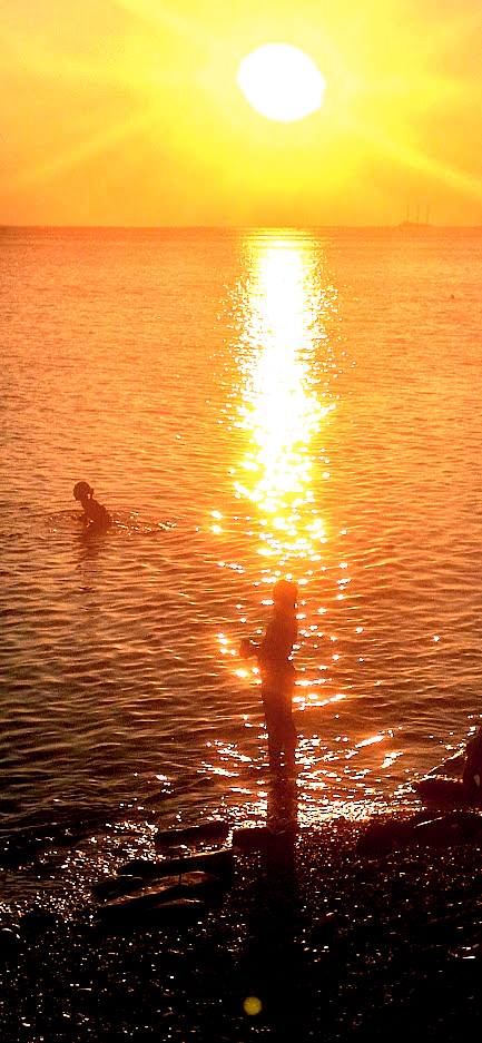 Spiaggia di sassi al tramonto. Il sole si riflette sulla superficie del mare e disegna le sagome di due ragazzine, una sul bagnasciuga, l'altra in acqua.