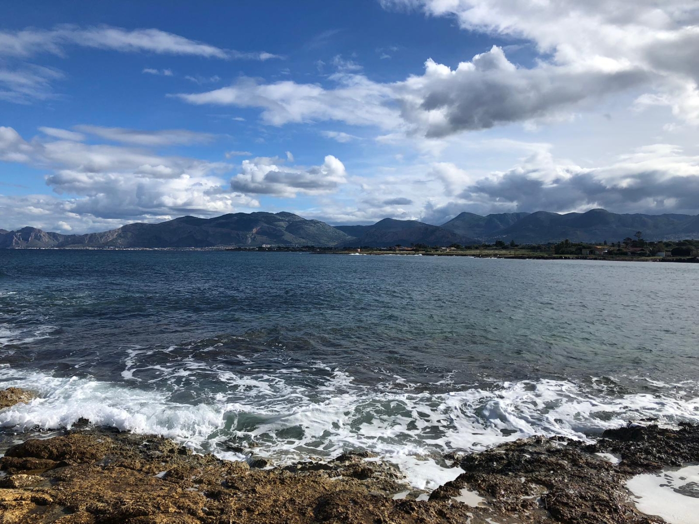Una giornata qualunque in quest'autunno novembrile, quando la natura dà il più bel spettacolo di sé stessa. Non più il cielo terso scolorito dall'afa e il mare immobile, non più i colori sbiaditi da un sole atroce che non abbronza ma brucia. Ora la luce è tagliente, accende i contrasti tra il blu profondo del mare e il giallo del calcare della scogliera; il cielo, denso di nuvole in corsa, disegna ombre su un mare risvegliato da un lungo sonno, che respira forte contro le rocce nell’attimo in cui un’onda si infrange alta sugli scogli, come se ogni onda volesse incidere una parola sul bordo delle pietre. A tratti il vento si alza, increspa la superficie dell’acqua; nelle pozze tra gli scogli resta una schiuma sottile, memoria effimera del passaggio delle onde. Più che semplici vedute, queste immagini sono punti di ascolto: invitano a sentire il rumore del mare, il fruscio del vento, il peso delle storie non dette che abitano ancora le pietre di questo che fu un ameno luogo, perché la fotografia è bugiarda, ti consegna solo l’imbeccata del fotografo. Basta spostare di poco lo sguardo e il quadro cambia. Alla mia destra la lunga striscia d’asfalto della bretella per l’aeroporto taglia il territorio con il suo flusso continuo di auto. Alle mie spalle il complesso della “Tonnara dell’Ursa” veglia su questo teatro naturale come un’antica sentinella di pietra, ponte silenzioso tra ieri e oggi. Dietro ancora, la fila di muri, recinzioni e case sul demanio continua a raccontare una storia di occupazioni, abusi e silenzi: la stessa che denunciammo con le mostre fotografiche “Mafia e territorio” insieme a #peppinoimpastato  a #radioaut  All’orizzonte la Punta del Raìs continua a vegliare ed esistere in filigrana, dietro il nome ufficiale dedicato ai giudici uccisi dalla mafia, a ricordare che la toponomastica, come la memoria, cambia ma non si cancella.
