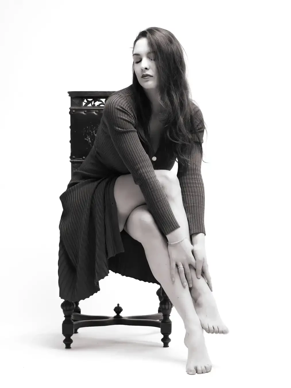 A woman with long hair sits barefoot on an ornate chair in a black-and-white photo, wearing a ribbed dress, eyes closed, one leg crossed over the other.
