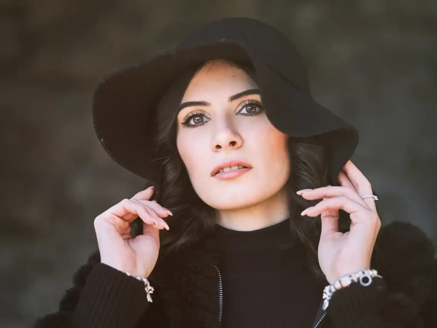 A woman with long dark hair wears a black wide-brimmed hat, black turtleneck, and black fuzzy jacket, holding the hat brim with both hands.