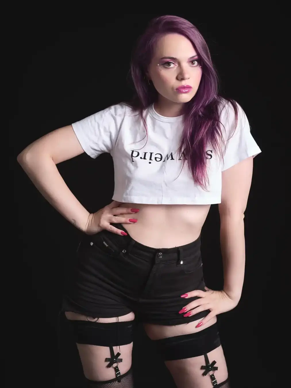 A person with purple hair poses against a black background, wearing a white cropped t-shirt with reversed “weird” text, black shorts, and black garter belts.