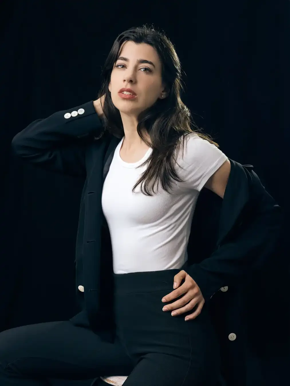 A woman with long dark hair poses in a white t-shirt, black pants, and black blazer against a dark background, one hand on her hip, the other behind her neck.