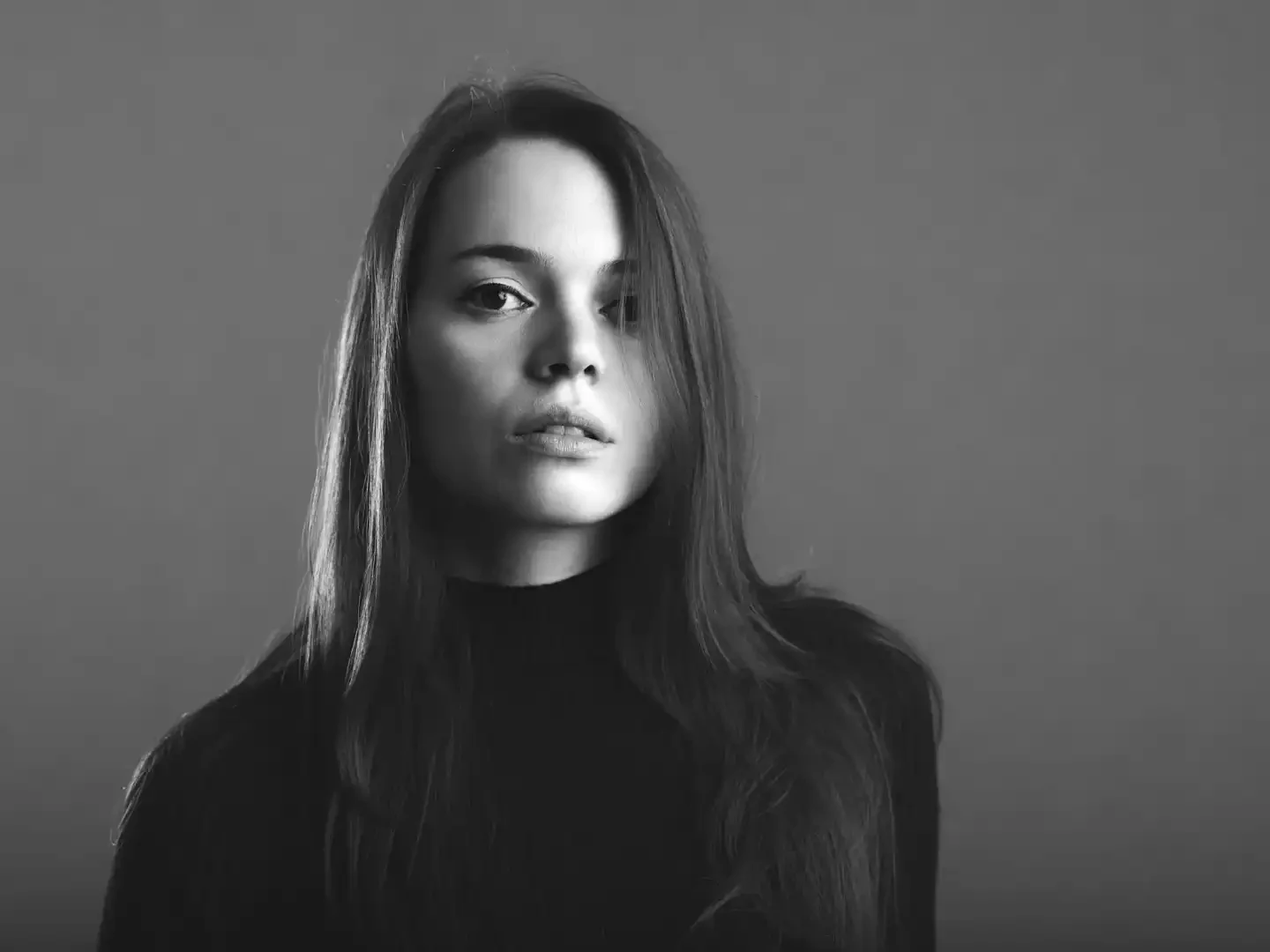 Black and white portrait of a woman with long hair wearing a dark turtleneck, against a plain gray background.