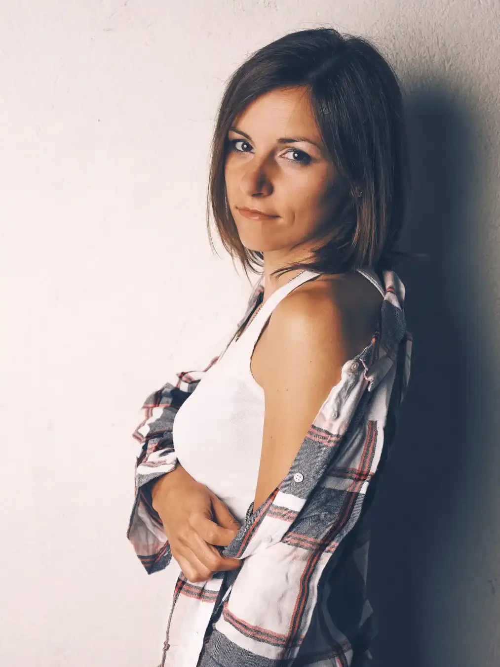 A woman with short brown hair wears a white tank top and an unbuttoned plaid shirt, standing against a light-colored wall.