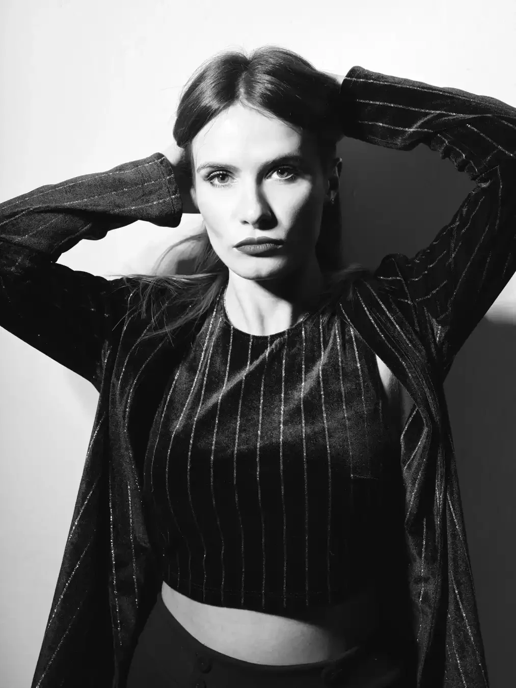 Black-and-white portrait of a woman with hands behind her head, wearing a pinstriped cropped top and jacket, gazing at the camera.