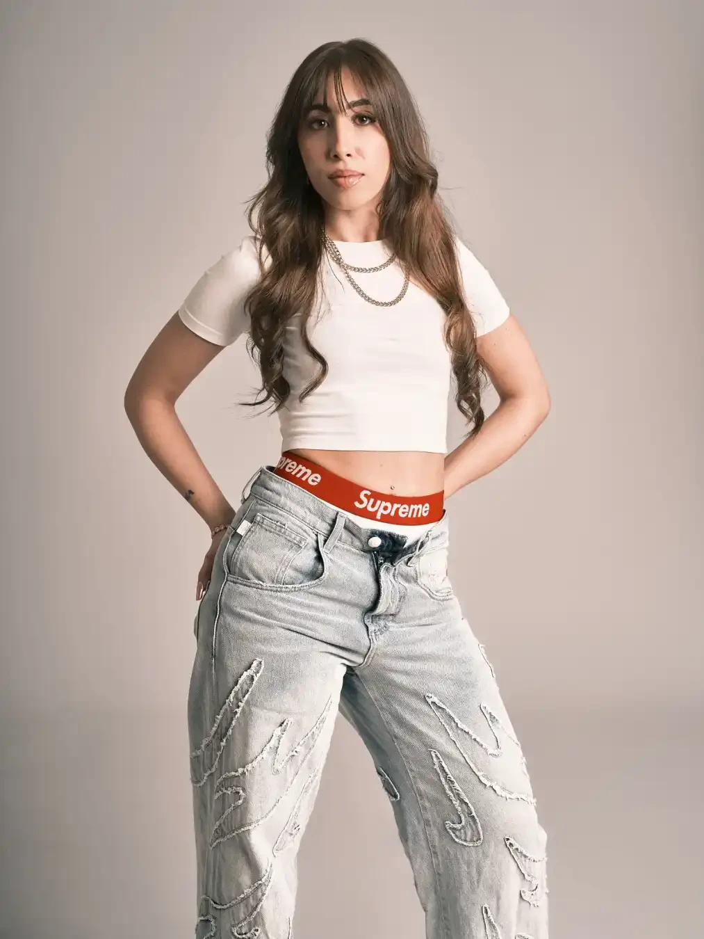 A woman with long wavy hair poses in a white crop top, light blue ripped jeans, and a red "Supreme" waistband against a plain background.
