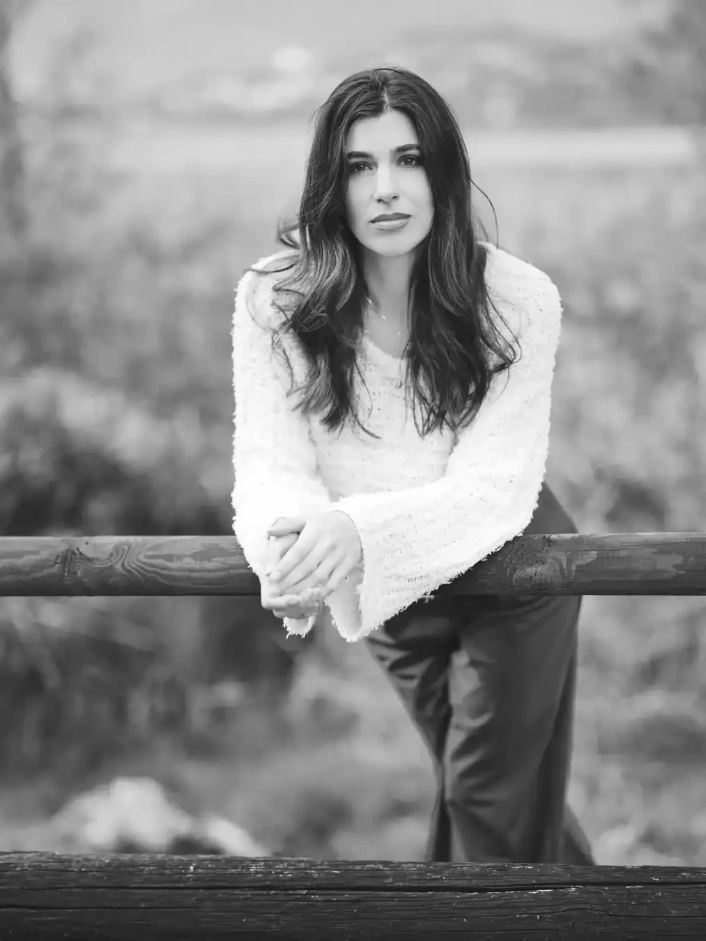 A woman with long hair in a textured sweater and pants leans on a wooden railing, hands clasped, in a black-and-white outdoor scene.